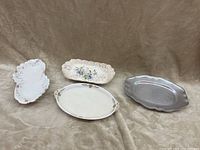 Four serving platters displayed on a fabric surface: three ceramic with decorative floral and embossed designs, one metal with patina and smooth finish.