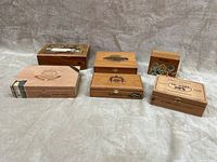 Six wooden cigar boxes arranged on a neutral background, showing different brand labels and artwork on each box.