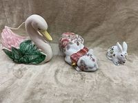 Three porcelain decorative animals on a beige surface: swan planter, reclining cat, and small rabbit figurine