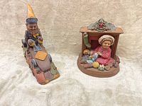 Two Tom Clark gnome figurines on a neutral fabric background, one seated on a log, the other inside a fireplace setting.