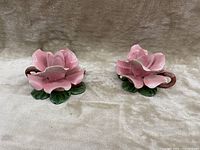 Pair of pink flower-shaped porcelain candle holders with green leaves base, shown side by side on beige fabric.