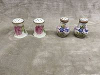 Pair of white porcelain salt and pepper shakers with pink flower decoration and metallic tops next to a pair of white and light purple porcelain shakers with purple flowers and scalloped tops.