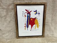Front view of whimsical signed painting in wooden frame on fabric background.