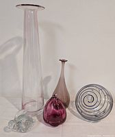 Photo showing five pieces of modern art glass including tall Scandinavian style violet vase, Quebec pink vase, clear hummingbird feeder, purple pumpkin fruit and clear rabbit figurine.