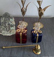 Photo showing the gilded tin bird candle holders, blown glass gift package candle holders in red and blue, and brass candle snuffer arranged on a grey fabric surface
