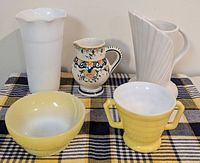 Five vintage items on plaid cloth: tall milk glass vase, white ceramic pitcher, colorful floral creamer, yellow Hazel Atlas creamer, yellow Fire-King bowl.