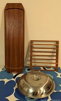 Three pieces shown together: rectangular teak trivet with horizontal slats, long teak tray, and stainless steel covered serving dish with handle.