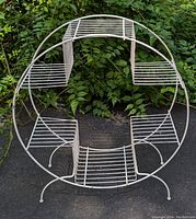 Photo front view of white wire plant stand showing circular frame and five shelves, outdoors with greenery background.