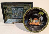 Rectangular Olinala lacquer tray with colorful traditional painted design and round black glaze Tlaquepaque plate with hand painted house and tree motif displayed together.