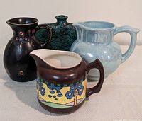 Vintage lot of four pottery pitchers, showing front and side angles. Includes a dark brown Pantry Parade pitcher with colorful tree pattern, green vessel, Gouda Holland signed piece, and blue Czechoslovakia pitcher.