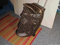 Brown monogrammed roller carry-on bag standing upright on a floor mat, showing the monogram pattern and shoulder straps