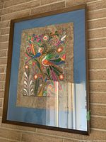 Front view of framed colorful bird print on bark-like texture mounted on blue mat and brown wooden frame hanging on brick wall.
