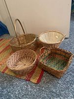 Four wicker baskets placed on a carpet with a beige door wall background; varying shapes and styles visible including handle basket and fabric lined basket.