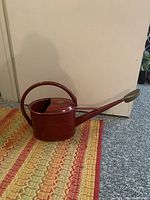 Full view of the red metal watering canister showing its shape, curved handle, and elongated spout with brass rose.