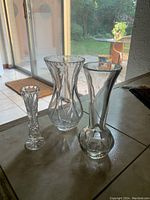 Overview photo showing three clear glass or crystal vases on a dark table by a window with garden background.