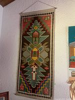 Full view of one colorful South American tapestry with geometric and face design hanging on a wall.
