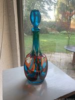 Photo of a tall glass decanter with a narrow neck and glass stopper, featuring an abstract stained-glass style pattern in multiple bright colors including blue, green, orange, red, and purple.