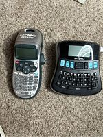 Photo showing two Dymo label makers side by side on a carpeted surface: one LetraTag handheld model and one electronic label maker with a larger keyboard.