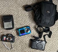 Image shows Canon G10, Pentax digital camera, small blue digital camera, DXG pouch, and Lowepro camera bag on carpet.