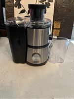 Side view of Dash fruit juicer showing stainless steel body, black plastic pulp collector on left, clear juice container on right, and cracked clear lid on top.