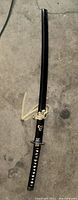 Katana in black scabbard with decorative cord tied, lying on concrete floor