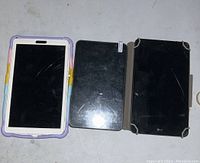 Photo showing three Android tablets side-by-side on a white surface. The left tablet has a colorful protective case and cracked screen. The middle tablet has a black screen with no visible brand or case. The right tablet is an LG tablet with a black rugged case and scratches on the screen.