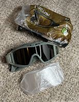 Photo showing military goggles with tinted lens, clear spare lens in plastic, and a branded hard shell case in plastic wrap on a carpeted surface.