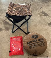 Photo of a camo folding stool, Mossy Oak Heat-A-Seat pouch, and red Skame Premium fire blanket pouch on concrete floor