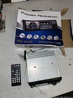 Photo showing the Power Acoustik box, car stereo receiver unit, and remote control on a surface.