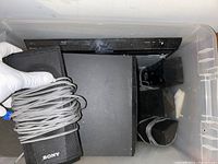 Samsung surround sound system components including the main unit and Sony Blu-ray player with power cords coiled, stored in a plastic bin.