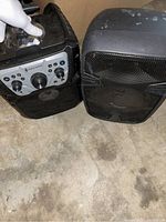 Photo showing two black portable speakers on a concrete floor. One speaker has a control panel with multiple knobs and ports, the other is bigger with a metal grille front. Both visibly scratched and used.
