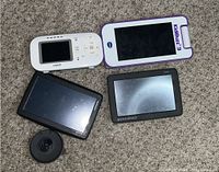 Four electronic devices on carpet: white Vtech Kidibuzz E tablet with purple trim, white Vtech GPS device, black Rand McNally GPS, and Garmin black round GPS mount. Devices show use, screens smudged.