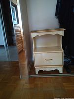 Photo showing a cream-colored wooden sidetable with gold trim, a glass top, one drawer with a gold handle, and a lower open shelf, placed next to a mirror and parquet floor.