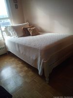 Photo of a white painted wooden single bed frame with headboard and footboard, dressed in white bedding and pillows, positioned near a window.
