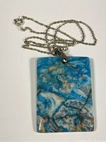 Photo showing full rectangular blue agate pendant with silver-toned bail attached to silver chain spread out, pendant facing up.