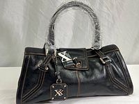 Front view of black leather handbag with two handles wrapped in plastic, Maxx New York tag attached, visible stitching and metal logo tag.