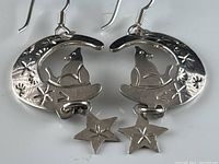 Pair of silver crescent moon earrings with sitting cat figures and dangling stars, showing engraved floral/star motifs and silver finish.