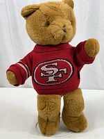 Front view of the light brown plush teddy bear standing upright wearing a maroon knit sweater with the San Francisco 49ers logo on the chest. Shows the sweater's sleeve striping detail and display stand at feet.