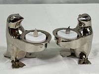 Front view of both polished metal penguin votive holders, each with battery-powered votive candles placed in the circular bowl each penguin holds.
