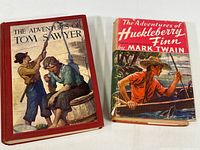 Two vintage Mark Twain children’s books side by side, The Adventures of Tom Sawyer (red hardcover) and The Adventures of Huckleberry Finn (worn dust jacket)