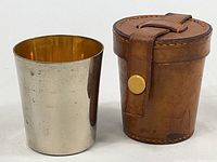 Silver-toned travel cup beside brown leather cylindrical carrying case with snap closure.