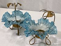 Pair of hand-formed blue ribbon glass dishes with ruffled edges on gold-colored engraved wire stands with leaf handle detail.