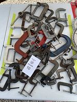 A pile of approximately 28 assorted C-clamps in different sizes and colors including silver, red, black, and rusted metal. Various handle types visible.