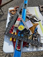 Top down view of assorted hand tools on a table outdoors, showing handsaws, levels, squares, folding ruler, tape measure, and gloves.