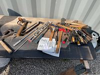 Wide shot of assorted hand tools laid out on a black surface with sunlight casting shadows, showing hammers, screwdrivers, levels, and a hand drill.