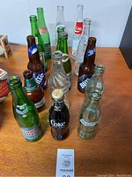 Overview of 15 vintage beverage bottles on wooden surface with some bottles containing residual liquid. Labels include Niagara Dry, Coke, Coca-Cola Classic, Labatt's Blue, Miller Lite, Coors, Bedford.