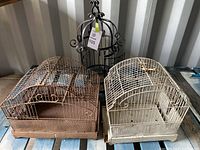Three different bird cages grouped together on a wooden surface, showing two rectangular cages and one decorative black cage.