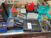 Overview of multiple containers and cases holding numerous drill bits of various sizes and types on a workbench.