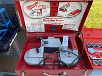 Open red metal carrying case showing Milwaukee portable band saw inside with black handle and adjustment lever, some wear visible, lid has printed Milwaukee branding and usage info.