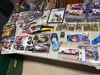 Wide view of table with NASCAR memorabilia including model kits, diecast cars, keychains, and magazines.